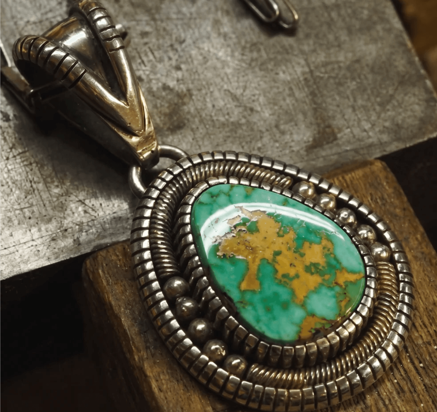 Handmade sterling silver pendant with a green turquoise cabochon, framed by coiled rope and bead wire, resting on the silversmith's workbench