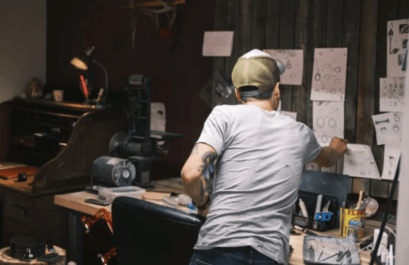 Christopher Bazan working at his studio bench, surrounded by pinned design sketches for ring and pendant commissions